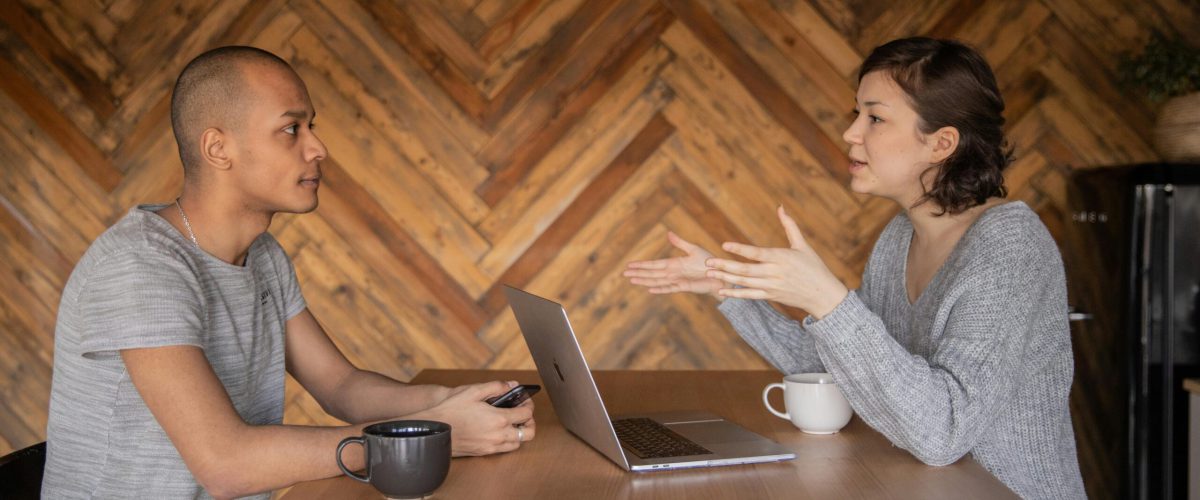 Focused woman explaining opinion to ethnic male coworker during business teamwork sitting at table with laptop and coffee cups in cozy kitchen against wooden wall and looking at each other